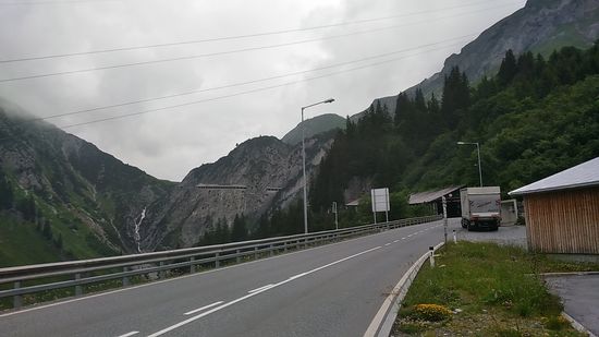 Hier an meiner Position gleich nach dem Abzweig von der Straße hinunter nach Bludenz hatte ich seit der Arlberg-Paßhöhe 4,7 km bergabwärts mit einem Verlust von gut 200 Höhenmetern hinter mich gebracht.
Jetzt stand dafür die Auffahrt zum Flexenpass ("Flexen" sind ein alter Begriff für die zahlreichen Kehren des früheren Saumweges an anderer Stelle) auf knapp 2,5 km und ca. 180 Höhenmetern bevor. 
Kurz nach dem LKW in der Seitenbucht erkennt man die Einfahrt in die sog. "Flexengalerie", eine Schutzgalerie vor Lawinen und Steinschlag, die sich mit kurzen Unterbrechungen bis ein kurzes Stück vor den Flexenpass an der Felswand hochzieht.
Die seit 1897 bestehende Flexengalerie musste übrigens von 2014 bis 2018 mit gesamten Bau- und Sanierungskosten in Höhe von 16,8 Mio. Euro zum Teil komplett neu errichtet werden. Dadurch wurde auch ein für die Zukunft angedachter "Erzbergtunnel" überflüssig, weil die sanierte Flexengalerie ebenfalls eine sichere Passage dieses Streckenabschnitts vor Steinschlag und Lawinen bietet.
Somit wurde mir rechtzeitig zu meiner getätigten Passage ein rundum ertüchtigter Fahrweg "serviert". Find ich gut !!!
In der linken Bildhälfte sieht man besonders bei Bildvergrößerung den weiß-schäumenden Flexenbach zu Boden stürzen.