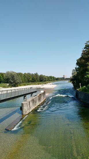 Im Hintergrund ist schon ein erstes Hochhaus zu erahnen.
Übrigens beginnt gleich hier anschließend rechterhand der Isar und des Radwegs das ausgedehnte Areal des Münchener Tierparks Hellabrunn.
Dieser Bereich an der Marienklausenbrücke mit dem links befindlichen Aussichtssteg stellt übrigens eine der Floßrutschen dar, über die die Ausflugsflöße in Richtung München hinunterrauschen.