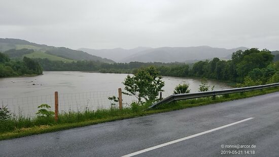 Aber wenige Kilometer weiter spricht die deutlich spiegelnde Straßenoberfläche wieder mal des Regens Sprache.
Blickrichtung ist hier übrigens ennsaufwärts, wobei die an dieser Stelle sehr breite Enns vom Ennskraftwerk Rosenau kurz vor der Ortschaft Garsten bedingt ist.
Das Kraftwerk selbst liegt in linker Bildrichtung, jedoch nicht festgehalten, da die Straße ab hier in linke Richtung etwas von der Enns wegführt und für mich im Rückblick nicht erkennbar war.
