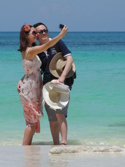 Zum Beispiel wie ein Pärchen mehrere Versuche unternimmt, sich vor der blaugrünen See zu porträtieren. Der Mann weiß gar nicht wohin mit den Hüten während seine Freundin fotografiert.