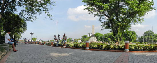 Obwohl es morgens um neun Uhr noch nicht so heiß ist, suchen sich die Leute lieber ein schattiges Plätzchen. Im Hintergrund ragt der Kilometer-Zero-Obelisk über die Bäume. Auf ihm sind die Entfernungen zu den wichtigsten philippinischen Städten notiert.