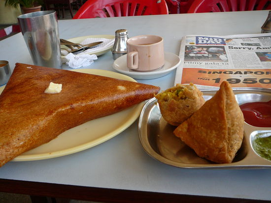 Mein erstes indisches Essen (links: Masala Dosa und rechts: Samosa). Vorher waescht man sich an einem Sammelbecken die Haende und dann isst man mit der Hand, wenn man moechte). Darueber: Chai, also Tee und eine Zeitung.