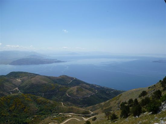 Wie ein langes Band zieht sich die Straßen den Berg hinauf. Dahinter ist die Meerenge zwischen Korfu und Albanien zu erkennen.