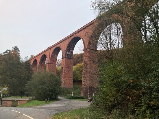 14 Hetzbach - Eisenbahnviadukt
Nachdem ich Amorbach in Richtung Kirchzell passiert hatte, war ich wieder mitten in einem meiner Wunschgebiete, im Odenwald. Seit ich zu Beginn meines beruflichen Unruhestandes 2013 eher zufällig hier die ersten Straßenkontakte knüpfte, war ich in jedem Jahr einige kurvige Tage in diesem Mittelgebirge unterwegs. Mit maximal 626,8 m ü NN gar nicht so wild, aber von einer Vielzahl herrlicher Kurvenstrecken durchzogen. Dazu gehört auch eine ganze Reihe von Bergrennstrecken, die jedes Jahr noch offiziell gleichmäßig oder auch schneller genutzt werden. Es gibt aber einen deutlichen Wermutstropfen, denn einige Abschnitte sind an den Wochenenden von Freitagabend bis Sonntag für Motorräder verboten. Der Platz für Gedenkkreuze wurde leider zu knapp.
Ich hatte mir für meine Ankunft den kurvigen Berg zwischen Ernsttal und Kailbach vorgenommen, den ich zum Einstimmen doppelt befahren habe. Dann ging es, dem römischen Limes folgend, nach Hesselbach und über eine einspurige, schmale Waldstraße hinunter nach Schöllenbach. Hier beginnt am bergseitigen Ortsausgang die Krähbergstrecke bis hoch zum Reußenkreuz und von der anderen Bergseite als Pendant von Hetzbach bis auch zum Reußenkreuz. In der dortigen ehemaligen Gaststätte hatte ich mehrere Jahre mein Quartier, war also auf sehr gut bekanntem Terrain. Heute befindet sich darin eine Jagdschule. Bei halbwegs passendem Wetter treffen sich Donnerstagabend viele Motorradfahrer, nicht nur zum Schwatzen. Wer möchte, kann bei YouTube z.B. unter Krähberg im Odenwald und dann in der Folge Videos finden. Ich fahre aber ohne so viel Gerede!-