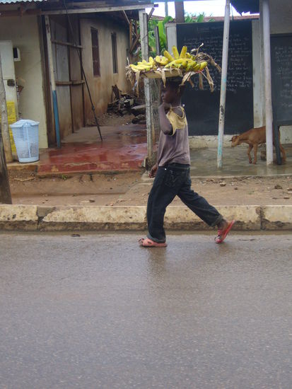 ein kleiner Junge trägt einen großen Korb mit Bananen auf dem Kopf.