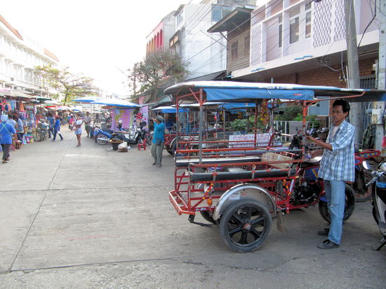Rechts im Bild ein Samlor. Hinter dem Fahrer kann man das drangeschraubte Mopped erkennen und den Sitz des Fahrers. Die linke Hälfte ist der Sitzbereich für Fahrgäste. Der Fahrer der davor steht, schaut grad in Fahrtrichtung. Wenn man mit dem Samlor über die Straßen heizt, dann fühlt man sich ein wenig wie in einer Schubkarre.