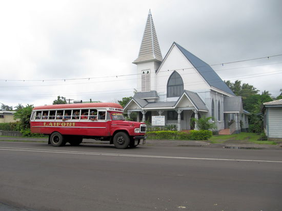 Ein typisches Samoabild! Auf Samoa haben die Missionare ganze Arbeit geleistet - hier wimmelt es nur so von Kirchen und selbst das kleinste Dorf hat noch eine Kirche. Der Bus im Vordergrund ist ein typisch samoanischer Bus. Es sind alte LKWs mit selbstgebauten Fahrgastbereichen aus Holz. Fenster gibt es keine, lediglich kleine Rollos, die man bei Regen runterlassen kann.