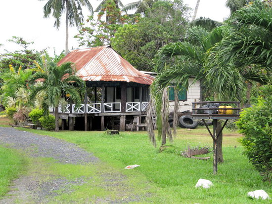 Ein samoanisches Wohnhaus mit dem traditionellen offenen Bereich (linke Seite) und einem geschlossen Bereich (rechte Seite), der mehr Privatsphäre bietet. Interessant ist der Kasten auf einem Pfahl vorne rechts im Bild. Es ist eine samoanische Mülltonne, die dafür sorgt, dass keine wilden Hunde oder anderen wilden Tiere vom Müll angezogen werden.