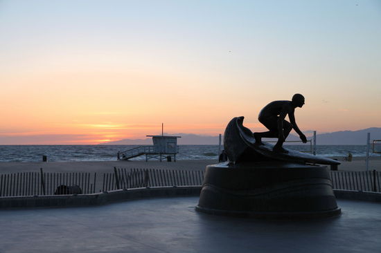 Eine Surferstatue am Hermosa Beach im Sonnenuntergang. Im Hintergrund eine der typischen Lebensretterhütten wie man sie aus Baywatch kennt.