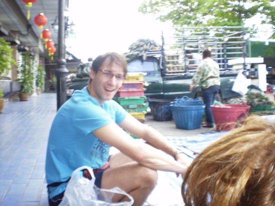 Das ist Gaspard. Noch gut gelaunt beim warten auf den Bus. Im Hintergrund ein typischer Marktstand in Thailand.