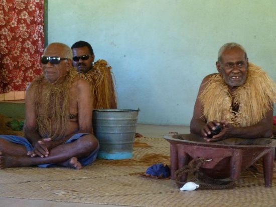 Rechts: der Chief, links: 2 der Ältesten. Ihr wisst noch was ich über das Sonnenbrillentragen gesagt habe?...
Der Chief sitzt üübrigens hinter einer Holzschale, die das Quasi-Nationalgetränk der Fijis enthält: Kava. Ein Pfefferwurzelextrakt, der mit Wasser vermischt und in einer Kokosschale gereicht wird. Schmeckt wie Pfützenwasser, sieht auch so aus und wirkt leicht bis mittelstark narkotisierend auf Lippen und Zunge. Eine Kavasession kann sich stundenlang hinziehen und resultiert in 30 Schalen Kava und einem tiefen Schlaf. Die Kavazeremonie ist aber auch eine Art Begrüßungsritual. Da mussten wir natürlich auch mitmachen. Wir Frauen saßen artig in einer Reihe hinter unseren Männern und nach bestimmten Verhaltensregeln nahme jeder einen Becher vom Schlammsüppchen. Es wurde gebetet und gesungen, dann war der Teil vorbei.
