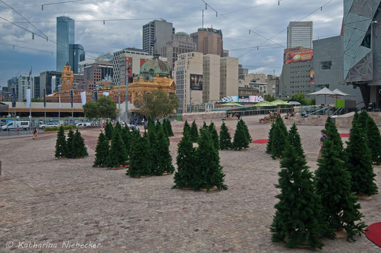 Ein Blick vom Federation Square über die kleinen Bäume auf das CBD mit der Flinders Street Station....