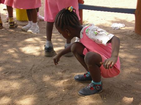 Eines der Kinder zeichnet im Sand auf dem Boden. Im Hintergrund sieht man ein Stückerl Klettergerüst.
