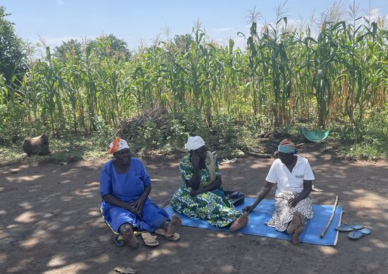 Die Damen sitzen wie beim letzten Mal im Schatten und klönen—Ich erfahre, dass zur Unterstützung junge Frauen aus dem Nachbardorf gekommen sind