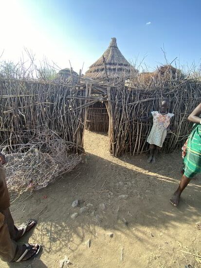 Hier stehen wir vor einem Zaun, der die Hütte(n) seiner Bewohner umkreist. Die Zäune sind aus Dornenbuschsträuchern oder -bäumen und soll Eindringlinge abhalten. Der Durchgang im Zaun ist niedrig, er wird von innen dann mit einem Dornenstrauch geschlossen, der von Außen nicht entfernt werden kann. So sind die Bewohner sicher. Auch die Kleintiere nimmt man mit hinein, dort sind sie geschützt. Innerhalb eines solchen Kreisels lebt eine Familie. Das sind teilweise mehrere Hütten.