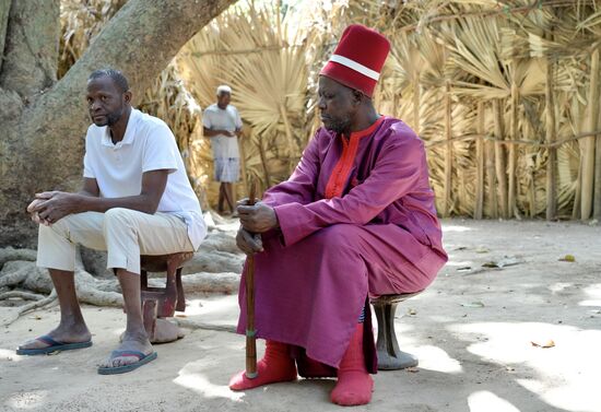 Voller Demut sitzt er auf einem Holzschemel aber als Zeichen seiner Erhabenheit läßt er seine Lakaien für sich sprechen. Er ist gekrönt mit einer Art Kochmütze in derselben Farbe wie sein schlichtes scharlachrotes Gewand und trägt anstelle eines Zepters einen Reisigbesen