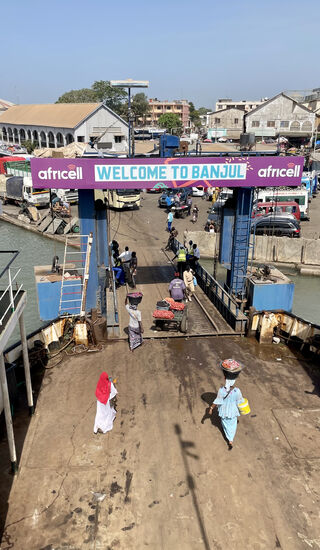 Irgendwie schafft es Dawda, dass wir als erste auf die Fähre kommen. Weit hinten, bei dem weißen großen Gebäude, gibt es einen riesigen Wartebereich, dort warten hunderte von Menschen dicht gedrängt