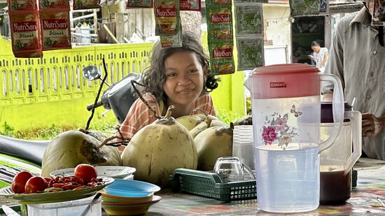 Diese junge Dame kommt mit ihrem Opa auf dem Motorbike vorbei und sie kaufen sich ein paar Getränke zum mitnehmen. Dazu wird eine der Tüten, die man oben hängen sieht in eine Plastiktüte mit heißem Wasser aufgelöst. Dann kommt kaltes Wasser dazu und schließlich Eiswürfel und ein Strohhalm. Tatsächlich gibt es hier noch Plastikstrohhalme  
Viele Getränke werden hier in Plastikbeutel, anstatt in Becher gefüllt