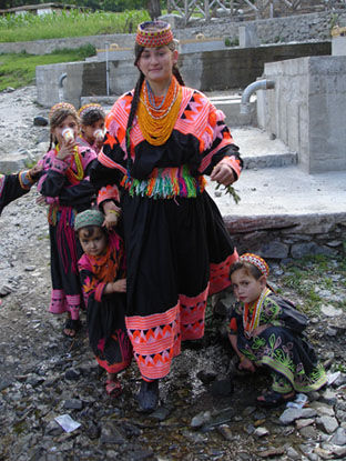 Alltagskleidung der Kalash-Frauen. Schwarz-bestickte Robe mit breitem Wollgürtel und auf dem Haar eine nach hinten herabfallende Schärpe, die völlig mit Perlen, Blutkorallen, Kaurimuscheln und Silber verziert ist. Sie tragen im Gegensatz zu anderen Gebieten in Pakistan keinen Schleier.
